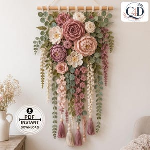 May include: A wall hanging featuring a collection of crocheted flowers and leaves in shades of dusty rose, cream, and sage green. The arrangement is suspended from a wooden dowel and includes cascading floral vines and decorative yarn tassels.