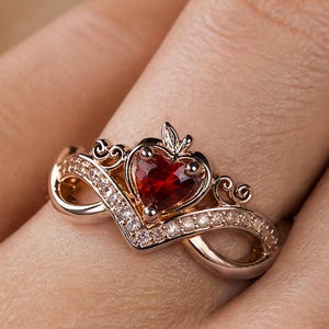 Op de afbeelding: Een ring in roségoudkleur met een centrale hartvormige rode edelsteen in een kroonachtig ontwerp. De band heeft de vorm van een oneindigheidssymbool en is bezet met kleine, heldere, fonkelende steentjes langs de bovenste randen van het metaal.