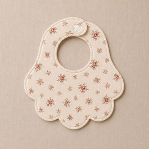 Peut inclure: Un bavoir pour bébé de couleur crème avec un bord festonné et un motif floral répétitif de petites roses roses et de feuilles vertes. Le bavoir est doté d'une fermeture à pression en plastique blanc au sommet de l'ouverture du cou.