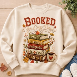 Peut inclure: Un sweat-shirt à col rond couleur crème avec une impression graphique représentant une pile de cinq livres aux motifs floraux brodés, une tasse fumante avec un cœur et des feuilles d'automne. Le texte indique BOOKED for Cozy Days.