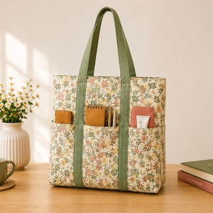 Peut inclure: Un sac fourre-tout rectangulaire en tissu crème orné d'un petit motif floral dans des tons de rose, jaune et vert. Le sac possède deux anses en tissu vert olive et trois poches extérieures à l'avant contenant un carnet, des stylos et un tube.