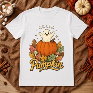 Peut inclure: Un t-shirt blanc à manches courtes orné d'un motif représentant un petit fantôme sortant d'une citrouille orange. Le dessin inclut des feuilles d'automne colorées, deux petits champignons et le texte "HELLO" au-dessus et "Pumpkin" en jaune.