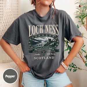 May include: A charcoal gray t-shirt featuring a graphic of the Loch Ness monster in a lake with a castle, surrounded by an oval border. The text reads LOCH NESS, Mystery of the Scottish Highlands, Timeless Wild Beauty, and SCOTLAND in white lettering.