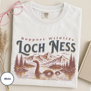 May include: A white t-shirt featuring a graphic print of the Loch Ness monster in a lake with mountains and pine trees. The text reads Support Wildlife LOCH NESS in dark blue and maroon. The shirt has a Comfort Colors brand label at the neck.