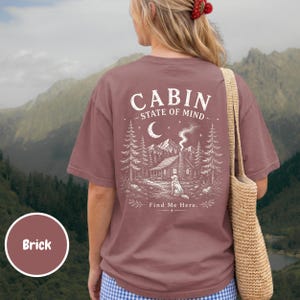 May include: A brick-colored t-shirt featuring a white graphic on the back. The design depicts a cabin in a forest with a dog, a crescent moon, and stars. The text reads "CABIN STATE OF MIND" at the top and "Find Me Here." at the bottom.