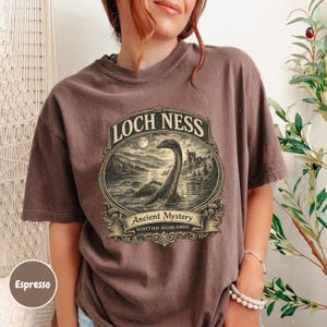 May include: An espresso brown short-sleeve t-shirt featuring a graphic print of the Loch Ness monster in a lake. The text reads LOCH NESS, Ancient Mystery, and SCOTTISH HIGHLANDS within an ornate, vintage-style oval frame design on the chest.