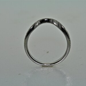 May include: A silver-colored ring with a curved design. The ring has a band with small, sparkling details. The ring is set against a plain, light-colored background. The ring's design is simple and elegant.