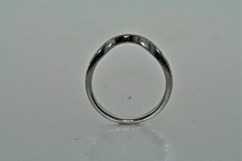 May include: A silver-colored ring with a curved design. The ring has a band with small, sparkling details. The ring is set against a plain, light-colored background. The ring's design is simple and elegant.