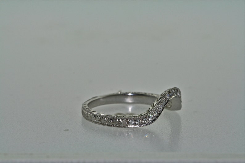 May include: A curved, silver-toned ring with a floral engraved band and a row of small, sparkling stones along the top curve. The ring is set against a plain, light background.
