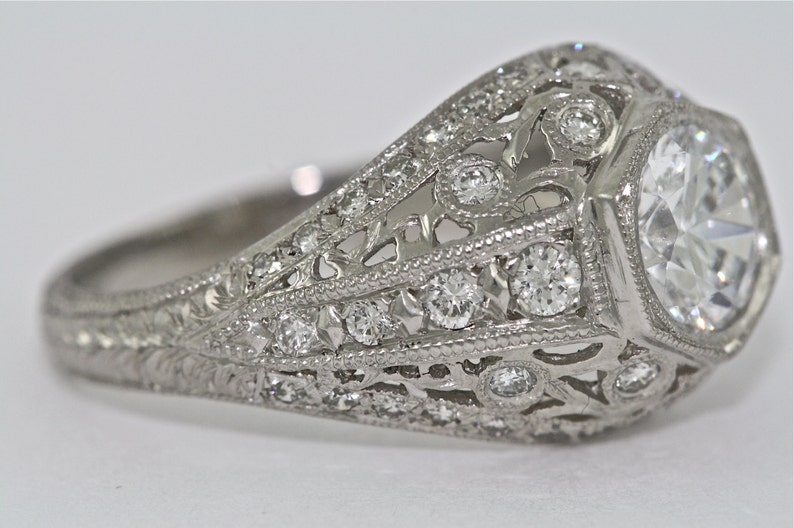 May include: A silver ring with a large, clear, octagon-shaped center stone surrounded by smaller diamonds. The band is intricately designed with filigree details and smaller diamonds.