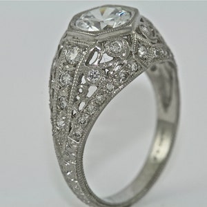 May include: A silver ring with a large, round, faceted diamond set in a filigree design. The ring is adorned with smaller diamonds around the center stone and along the band.