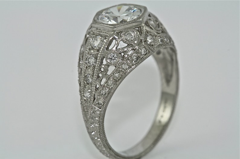 May include: A silver ring with a large, round, faceted diamond set in a filigree design. The ring is adorned with smaller diamonds around the center stone and along the band.