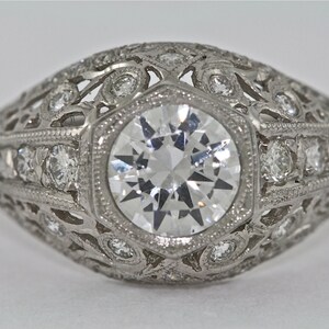 May include: A vintage-style engagement ring with a large, round, clear diamond center stone surrounded by smaller diamonds in a delicate filigree setting.