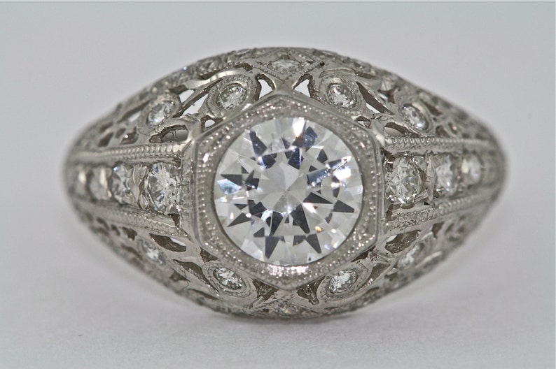 May include: A vintage-style engagement ring with a large, round, clear diamond center stone surrounded by smaller diamonds in a delicate filigree setting.