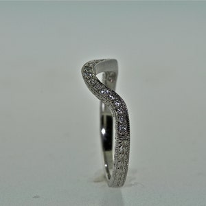 May include: A curved, silver-toned ring with a wave-like design. The top of the ring is encrusted with small, clear gemstones. The band features an engraved floral pattern. The ring is set against a plain, light background.