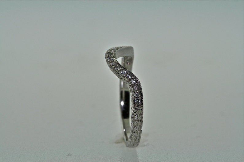 May include: A curved, silver-toned ring with a wave-like design. The top of the ring is encrusted with small, clear gemstones. The band features an engraved floral pattern. The ring is set against a plain, light background.