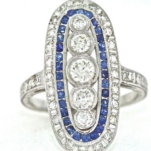 May include: A silver ring with a row of three round diamonds set in a oval-shaped frame. The frame is outlined with a row of blue sapphires.