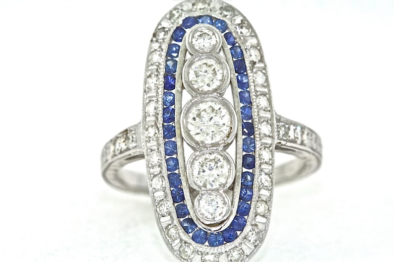 May include: A silver ring with a row of three round diamonds set in a oval-shaped frame. The frame is outlined with a row of blue sapphires.
