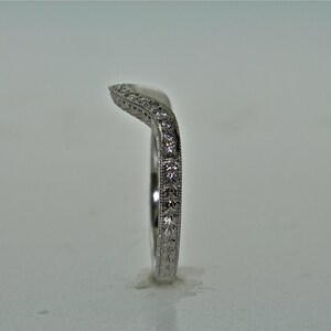 May include: A curved, silver-colored ring with a delicate design. The ring features small, sparkling stones and intricate patterns along the band. The ring is set against a plain background, highlighting its elegant details.