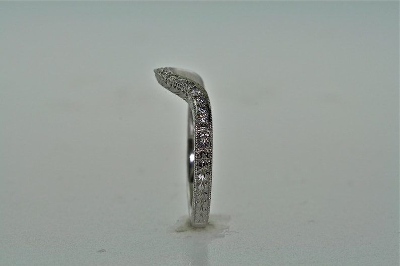 May include: A curved, silver-colored ring with a delicate design. The ring features small, sparkling stones and intricate patterns along the band. The ring is set against a plain background, highlighting its elegant details.