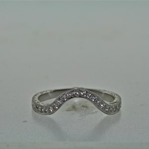 May include: A curved, silver-toned ring with a row of small, clear gemstones set along the top curve. The band features a detailed, textured pattern. The ring is designed to curve around another ring.