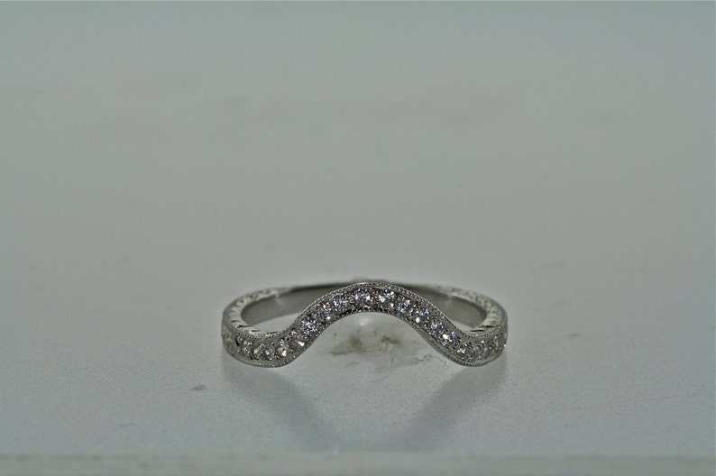 May include: A curved, silver-toned ring with a row of small, clear gemstones set along the top curve. The band features a detailed, textured pattern. The ring is designed to curve around another ring.