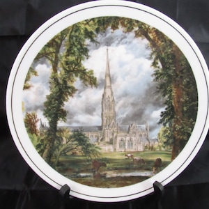 May include: A white ceramic plate with a gold rim features a painting of a cathedral with a tall spire. The cathedral is surrounded by trees and a small stream. The sky is cloudy and gray.