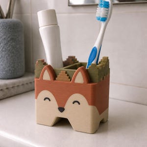 May include: A fox-shaped toothbrush holder with a matte finish, featuring a rust-orange and cream face with black eyes and a nose. The top edge has a green, blocky, castle-like texture. It holds a white toothpaste tube and a blue and white toothbrush.