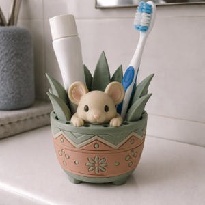 Puede incluir: Una figura de ratón beige asomándose desde un soporte para cepillos de dientes de color verde salvia y terracota. El soporte presenta un borde estampado con formas geométricas y motivos florales, conteniendo un tubo de pasta dental blanco.