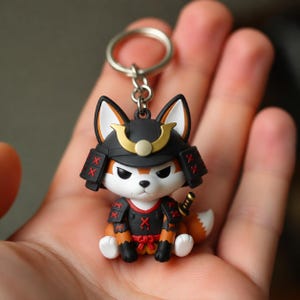 Può includere: Un portachiavi raffigurante un piccolo personaggio volpe stilizzato che indossa un elmo da samurai nero e oro e un'armatura nera abbinata con dettagli a punto croce rossi. La volpe ha il pelo bianco e arancione, orecchie a punta e una spada.