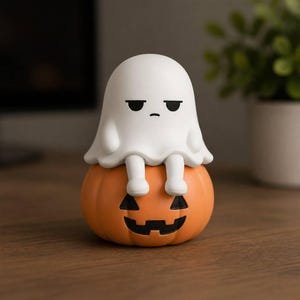 May include: A white ghost figurine with a bored facial expression sitting on top of an orange pumpkin. The pumpkin features a carved black jack-o'-lantern face with triangular eyes and a jagged mouth. The ghost has rounded arms and legs.