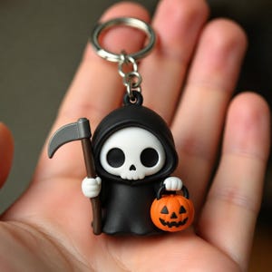 May include: A keychain featuring a small, stylized grim reaper figure wearing a black hooded robe. The figure has a white skull face, holds a brown-handled scythe in one hand, and carries a small orange jack-o'-lantern bucket in the other.