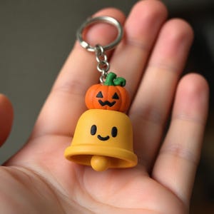 May include: A keychain featuring a yellow bell-shaped charm with a simple smiling face and a small orange jack-o'-lantern pumpkin figure attached to the top. The charm is connected to a silver-toned metal split ring by a short chain link.