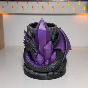 May include: A cylindrical desk organizer featuring a sculpted black dragon with purple wings coiled around a cluster of pointed purple crystals. The base and the upper rim of the container have a textured, rocky appearance in shades of dark gray.