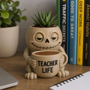Puede incluir: Una maceta de color beige con forma de calavera y expresión sonriente, que sostiene una taza beige a juego con el texto negro "TEACHER LIFE" impreso en el frente. Una planta suculenta verde está plantada en la parte superior de la calavera.