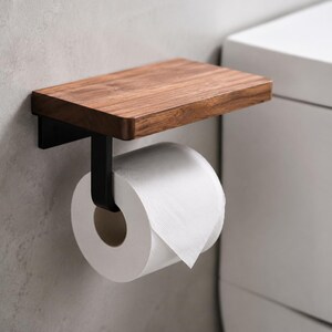 May include: A wall-mounted toilet paper holder featuring a rectangular dark brown wood shelf attached to a matte black metal bracket. The holder supports a single roll of white toilet paper, providing a functional storage solution for bathrooms.