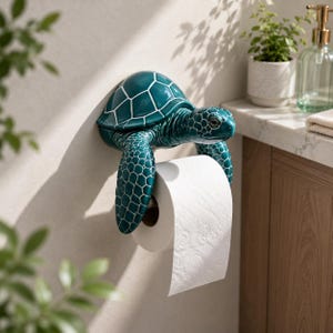 Op de afbeelding: Een wandgemonteerde toiletrolhouder in de vorm van een zeeschildpad met een glanzende blauwgroene afwerking en witte schildpatronen. De voorste flippers van de schildpad steken naar beneden om een standaard rol wit toiletpapier te ondersteunen.