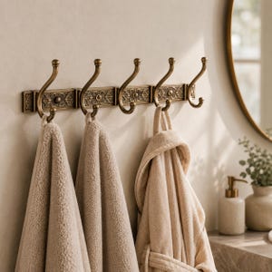 Peut inclure: Un porte-manteau mural composé de cinq crochets en laiton antique avec des plaques rectangulaires ornées. Deux serviettes beiges texturées et un peignoir beige assorti sont suspendus aux crochets, qui présentent des extrémités courbées.