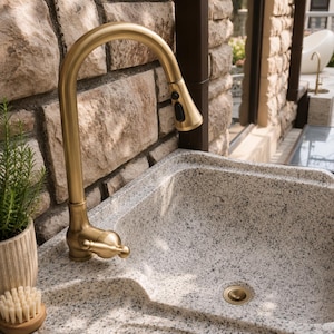May include: A brushed gold kitchen faucet with a pull-down sprayer head and a single lever handle, mounted on a speckled light gray granite sink basin. The faucet features a high-arc spout and a black button control for the sprayer function.