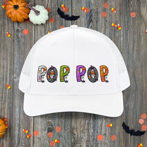 May include: A white trucker hat featuring the text POP POP in colorful, patterned letters. Each letter is decorated with Halloween motifs including pumpkins, spider webs, spiders, and candy corn. The hat has a solid front panel and mesh back.