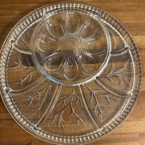 May include: A clear glass serving platter featuring a central circular section with an embossed shell pattern surrounded by a larger outer ring divided into four segments, each decorated with a raised branch motif and a textured beaded rim.
