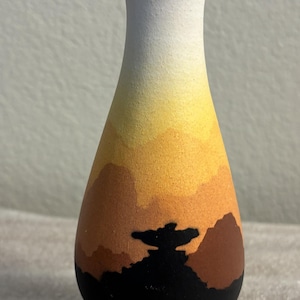 May include: A small ceramic vase with a matte finish featuring a gradient design that transitions from a white rim down through yellow, orange, and brown layers to a solid black base, with a black silhouette of a figure centered on the front.