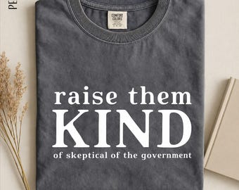 May include: A charcoal gray crew neck t-shirt with white text that reads "raise them KIND of skeptical of the government". The shirt features a Comfort Colors brand label at the inner neckline and is labeled as the color pepper.