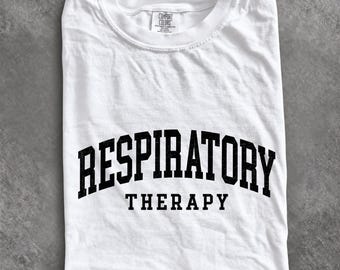 May include: A white short-sleeved t-shirt featuring the black text RESPIRATORY THERAPY in a bold, arched collegiate font across the chest. The garment has a crew neckline and a Comfort Colors brand label visible at the inner collar.