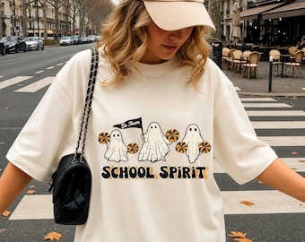 May include: A cream-colored short-sleeve t-shirt featuring a graphic of three white ghosts as cheerleaders holding orange and black pom-poms. One ghost holds a flag that says Go Team. The text SCHOOL SPIRIT is printed below in black.