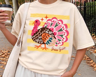 May include: An ivory short-sleeve t-shirt featuring a graphic of a turkey with a pink, red, and orange tail. The design includes yellow horizontal brush strokes, two small orange stars, and the text Gobble Gobble in cursive script.