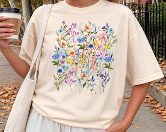 May include: An ivory short-sleeved t-shirt featuring a central graphic print of various colorful wildflowers including blue, yellow, orange, and purple blossoms with green stems and leaves. The shirt has a crew neckline and a relaxed fit.