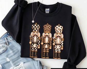 May include: A black crewneck sweatshirt featuring a graphic of three nutcrackers. The left nutcracker has a black and white checkered pattern, the middle has a leopard print, and the right has a black and white polka dot pattern.