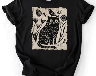 May include: A black short-sleeve t-shirt featuring a cream-colored rectangular graphic print of a black cat sitting among stylized flowers and a bird. The cat has wide eyes and is positioned in the center of the design with a bird on its head.