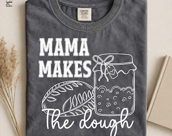 May include: A charcoal gray crew neck t-shirt featuring white line art of a loaf of bread and a jar with a heart tag. The text reads MAMA MAKES The dough. The shirt has a Comfort Colors label at the inner neckline and the color name Pepper.
